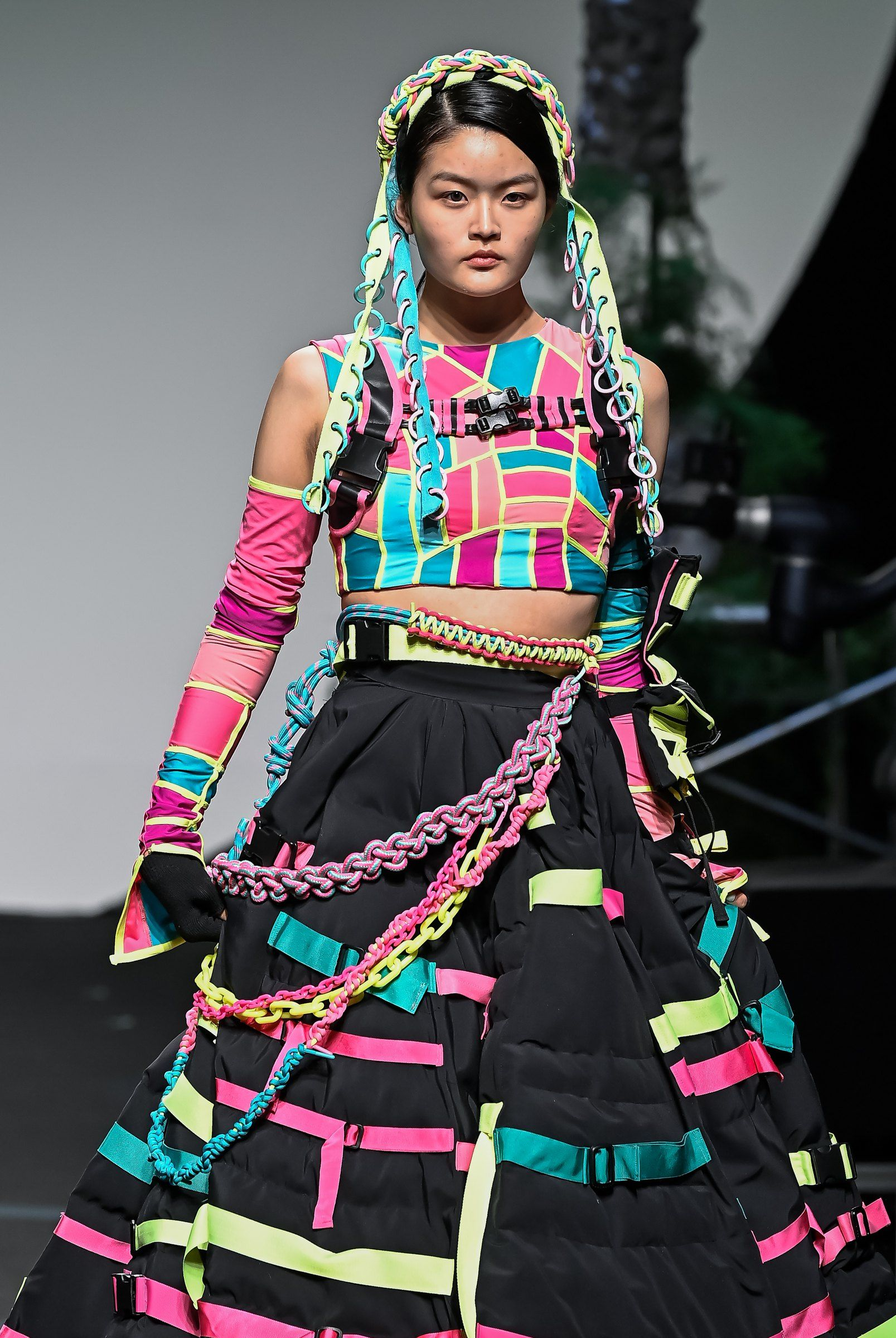 Model on the runway in a patchwork lycra crop top, hooded braid and a black quilted ball skirt striped with lime, pink and cyan tape
